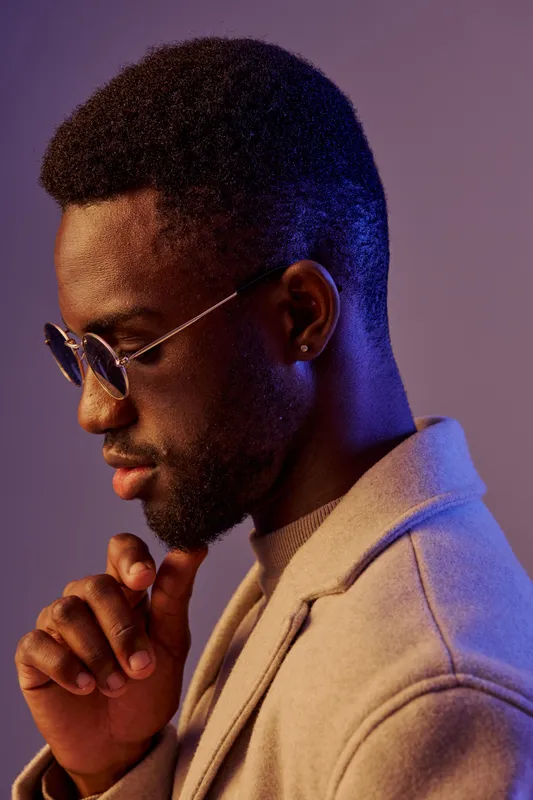 Thoughtful young African man in stylish sunglasses and coat, with a contemplative expression, against a vibrant purple background