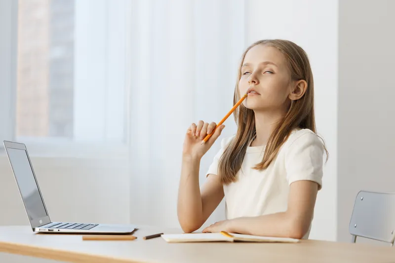 Thoughtful girl with a pencil, contemplating her studies at home, wearing a white shirt, with a laptop on the desk and bright, minimalistic background