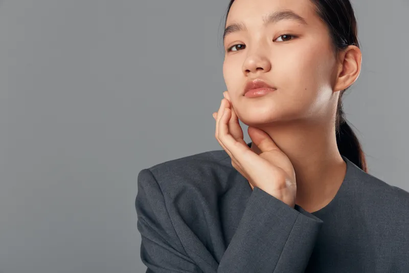 Young woman with clean skin and natural makeup posing with hand on face wearing gray jacket against plain background for beauty and skincare concept.