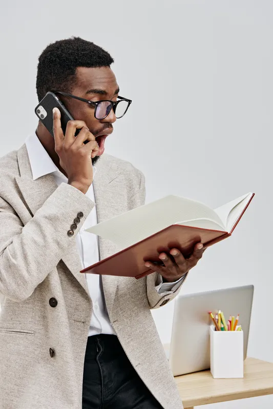 Professional young man of African descent with glasses, wearing a beige blazer and white shirt, talking on smartphone while reading a notebook, in a modern office setting. Business communication conce