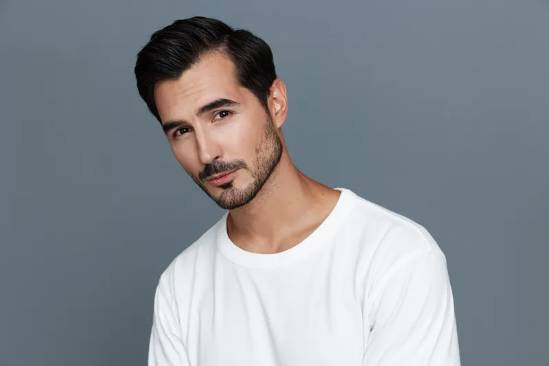 Handsome man with a well-groomed beard poses for the camera, wearing a casual white t-shirt against a soft gray background. Portrait photography concept.