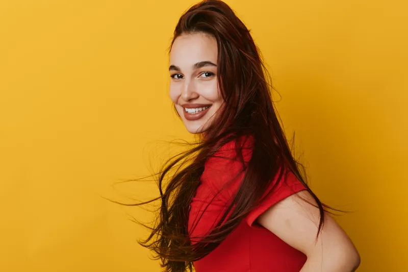 Young woman with long hair smiling and wearing red shirt posing against yellow background. Portrait of happy female with natural makeup and confident expression in studio lighting.