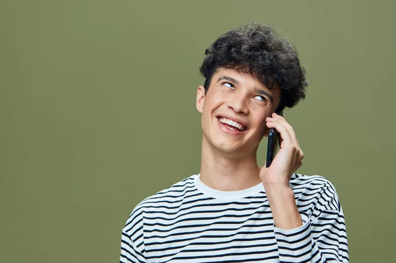 Young man talking on phone smiling, wearing striped shirt with curly hair, casual and cheerful expression on green background, communication and happiness concept.