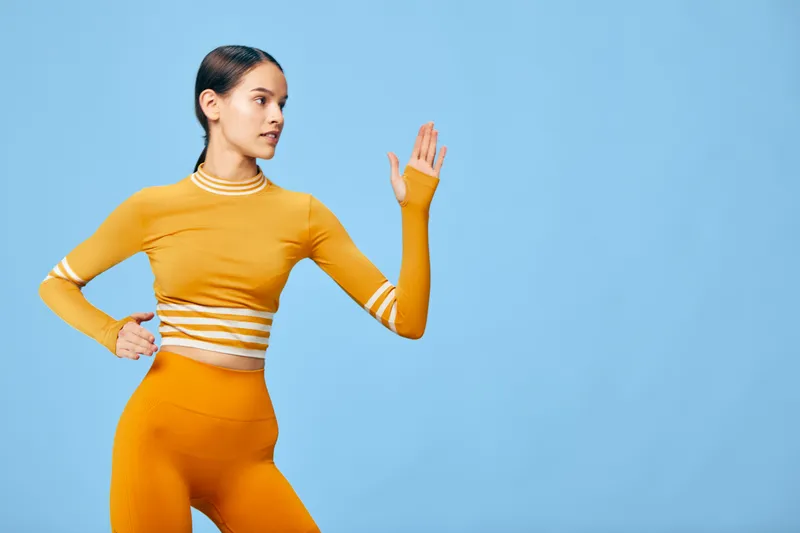 Young athletic woman in orange fitness outfit poses in a dynamic stretch against a clean soft blue background, highlighting sport posture, active lifestyle and modern workout fashion.