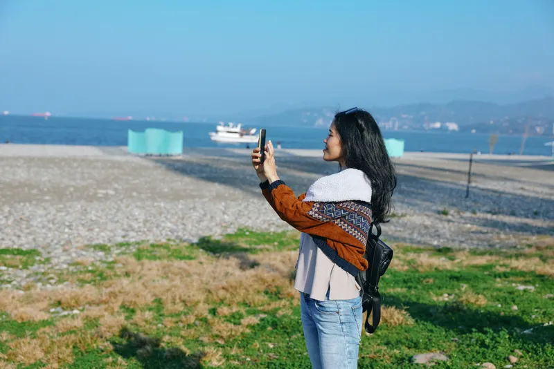 Selfie of a woman on a sunny beach, holding a smartphone to capture the sea, sand and distant shoreline, casual style with a knitted shawl and jeans, outdoor moment under bright light
