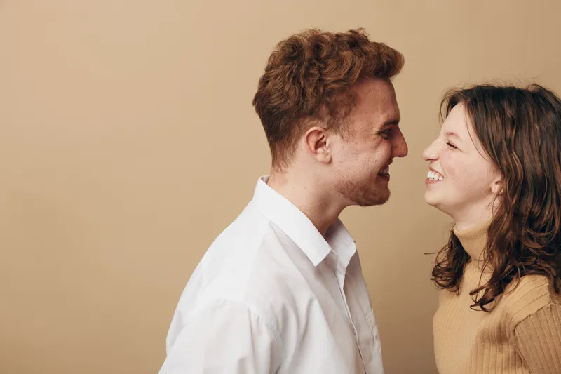 Smiling young couple facing each other closely, expressing happiness and affection. Casual clothing and warm beige background create a cozy and intimate atmosphere.