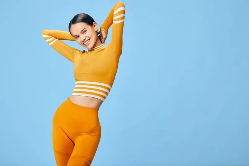 Fit young woman in bright orange workout set poses against a clean blue studio background, stretching her arms behind her head while smiling, showing athletic posture and sport.