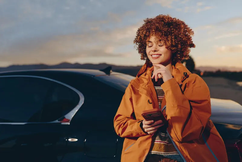 Woman with curly hair wearing orange jacket, standing near a parked car at sunset, smiling and holding a smartphone, casual woman lifestyle, outdoor travel mood, warm evening light