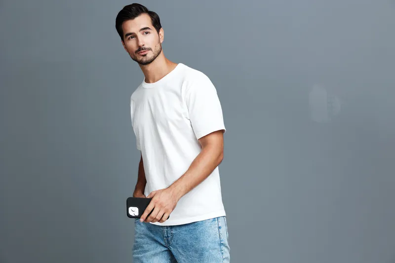 Stylish young man in a white t-shirt holding a mobile phone, looking confidently at the camera against a solid gray background. Fashion and lifestyle concept.