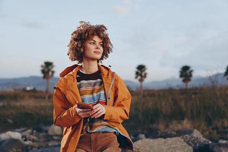 Woman outdoors lifestyle moment, curly hair, orange jacket, holding a mug, relaxed smile, soft daylight, scenic landscape with rocks and distant hills, natural colors open sky, dry field, calm