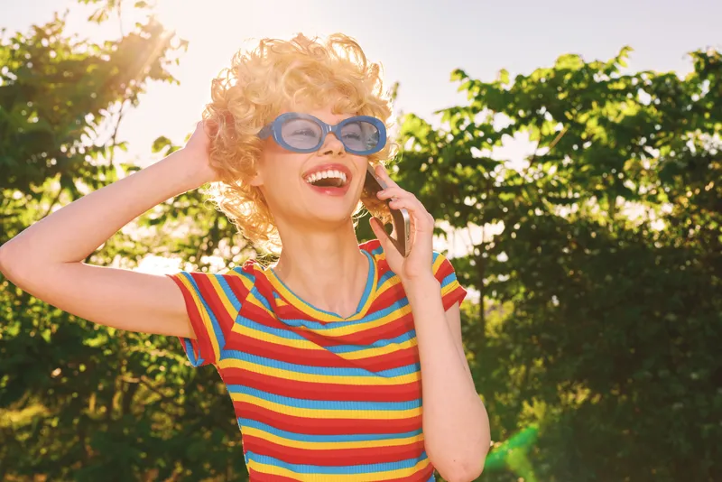 Young woman with curly blonde hair in a rainbow striped shirt talks on a mobile phone outdoors, smiling brightly in warm sunlight and enjoying a sunny moment during a casual outdoor shoot