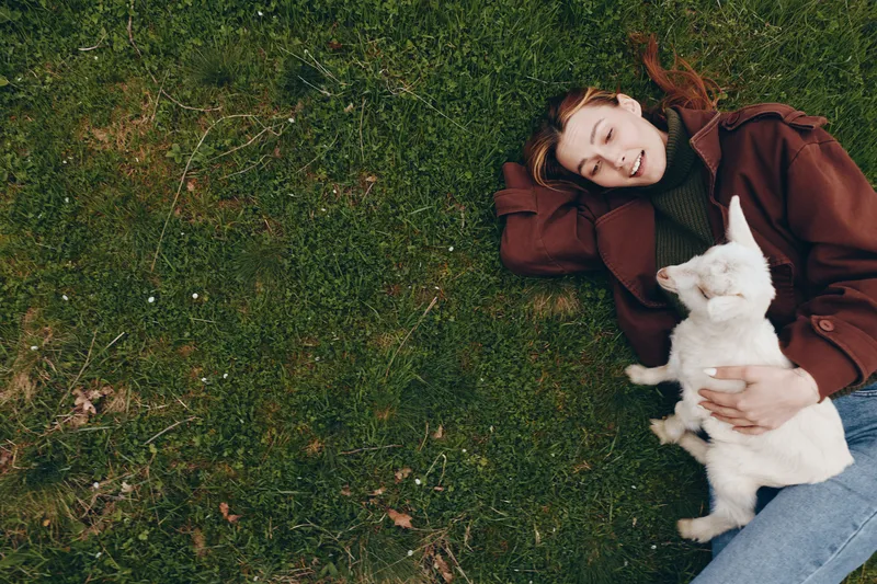 Relaxed woman lying on a grassy lawn, cuddling a white dog, enjoying outdoor leisure and tranquil companionship, a cozy lifestyle moment captured in natural light.