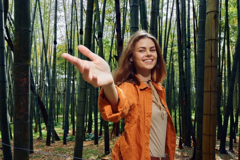 Woman in bamboo forest smiling and reaching hand toward camera, portrait outdoors in nature wearing orange jacket, relaxed expression among tall green bamboo stalks and trees.
