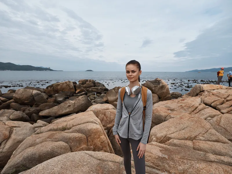 Young woman in activewear standing on rocky beach, enjoying nature with headphones, peaceful ocean view, symbolizes fitness and relaxation outdoors