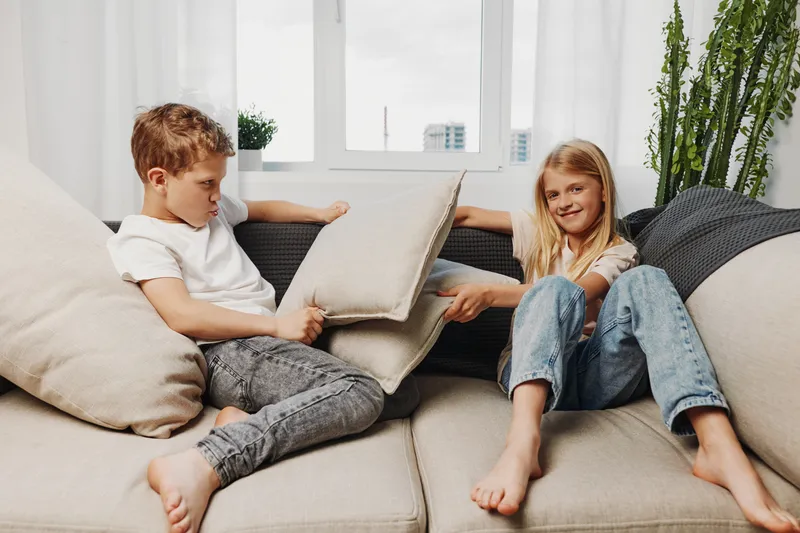 Two children playfully fight with cushions on a beige sofa in a bright living room with large windows and green plants. Concept of childhood fun, family moments, and home comfort.