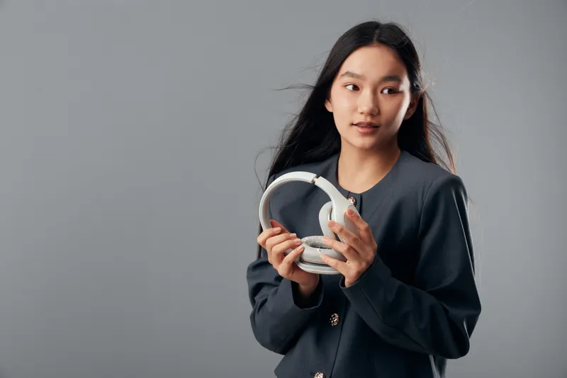 Young woman holding white headphones and looking aside, wearing dark jacket with long hair loose, studio portrait with neutral background for technology and lifestyle.