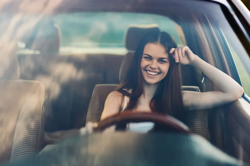 Happy young woman driving a car, smiling confidently, showcasing freedom and adventure on a sunny day. Bright, cheerful mood with a blurred landscape background.