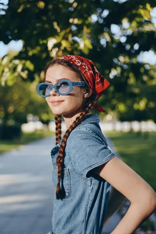 Young woman with braids wearing blue sunglasses and a red bandana smiling outdoors in a sunny park with green trees and a casual denim shirt.