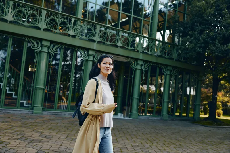 Woman with backpack walking in a park beside an ornate green fence, casual coat and jeans, daytime stroll, urban exploration, candid lifestyle moment, peaceful and purposeful