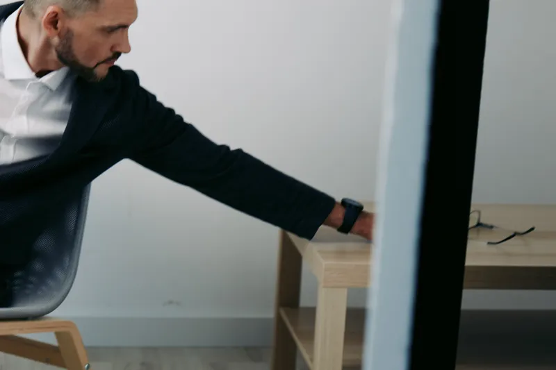 A focused business professional reaches toward a desk in a minimal, isolated color background, conveying determination, concentration, and poised readiness in a modern workspace.