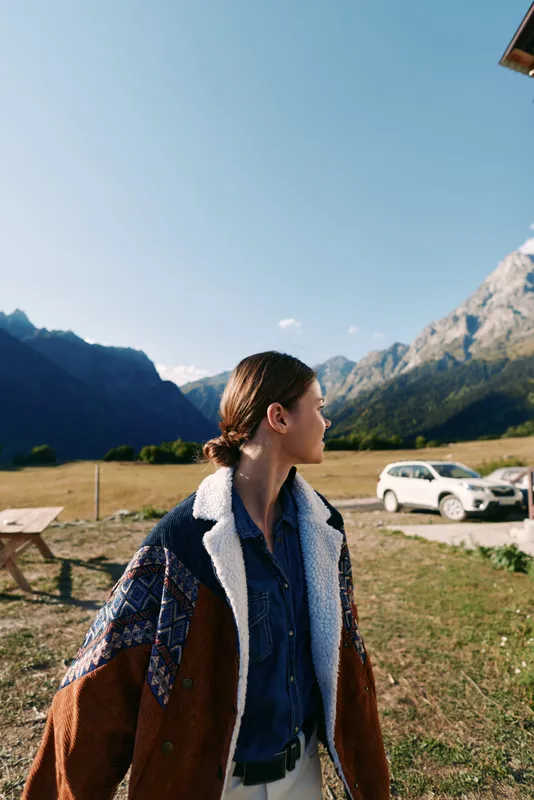 Woman in a warm jacket standing in a meadow by a white car, looking toward distant mountains during travel. Scenic outdoor portrait of nature, adventure and countryside road trip.