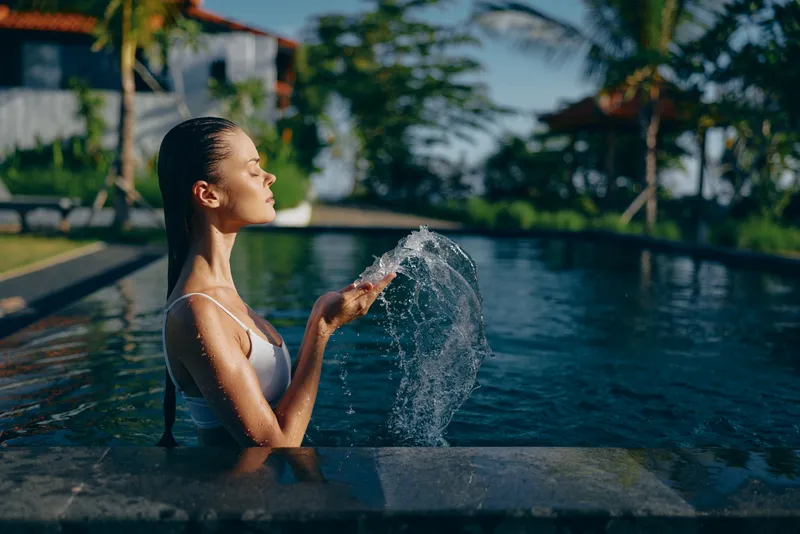 Woman in pool outdoors, sunlight on her face, relaxation and leisure in a summertime resort setting, water spray arcing softly, poolside calm and refreshing moment