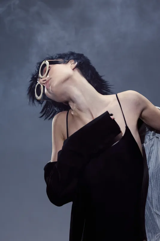 Stylish woman with short hair and sunglasses posing in black outfit with moody lighting and blurred background.