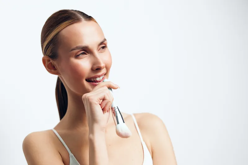 A smiling young woman with tied hair holds a makeup brush near her lips, looking away. The bright studio background highlights her natural skin and relaxed mood.
