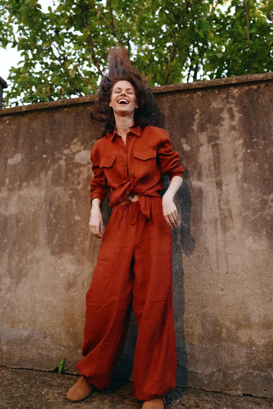 Joyful young woman in a red jumpsuit leaps beside a weathered wall, capturing a bold street style moment with bright energy and carefree charm