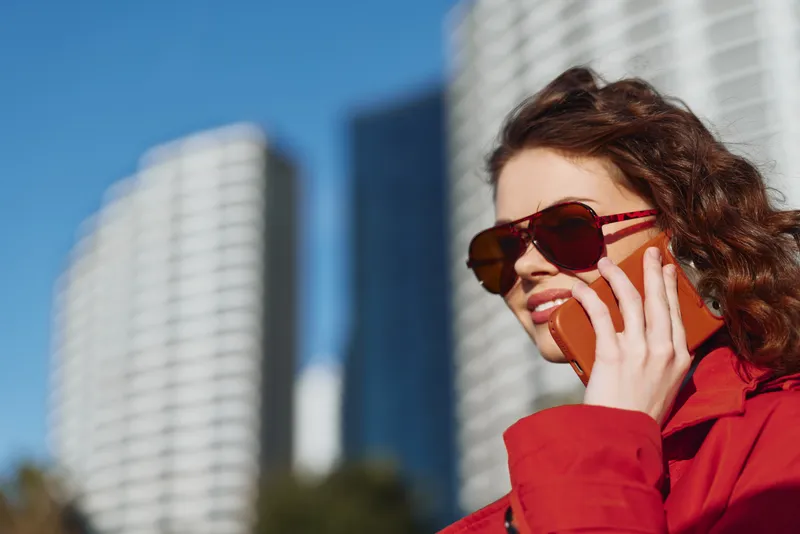Young woman with curly hair wearing sunglasses and a red jacket talks on a smartphone outdoors in an urban setting on a sunny day.