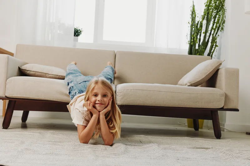 Happy blonde girl lies on carpet in bright living room with beige sofa and green plant. Cozy home interior and relaxed child concept.