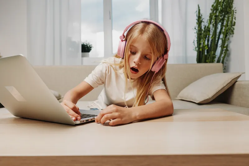Young girl with pink headphones using laptop on wooden table in bright room. She looks surprised and engaged, concept of technology, childhood, learning and digital interaction.