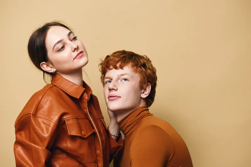 Two young people posing in stylish brown outfits against beige background. Woman wears leather jacket, man has curly hair and turtleneck sweater for fashion concept.