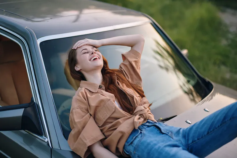 Young woman relaxing on a vintage car hood, smiling with joy, wearing a stylish casual outfit amidst a lush green background, emphasizing a carefree summer vibe.