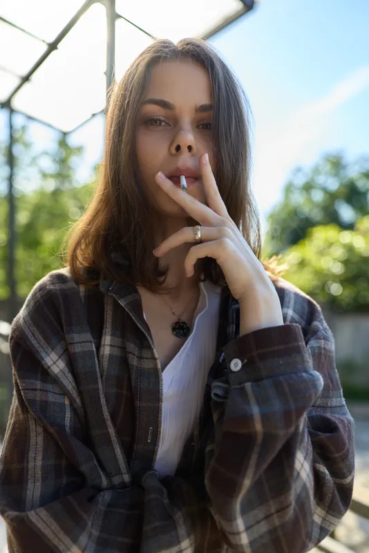 Woman with long hair wearing a plaid shirt smoking a cigarette outdoors. Casual lifestyle portrait, natural light, relaxed mood, candid moment, urban vibe, bright daylight, soft background blur