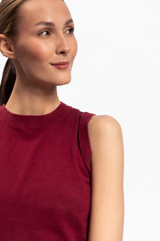 Young woman with clear skin and tied hair wearing sleeveless burgundy top looks aside with gentle smile. Bright studio background highlights natural beauty and casual style.