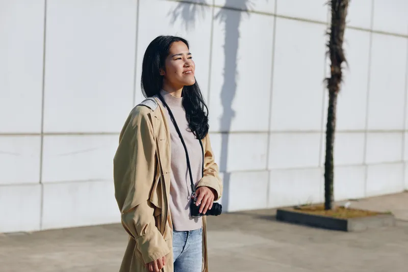 Woman outdoors in casual fashion, wearing a light coat and denim, standing beside a white wall in warm sunlight, relaxed street style and confident expression, urban backdrop and candid mood.