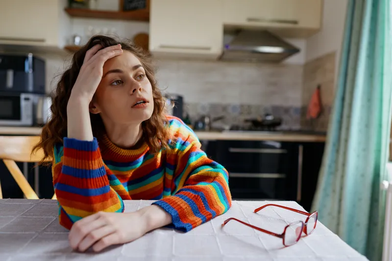A vivid striped sweater worn by a young woman sits at a kitchen table, one person visible, with a pensive bored expression in a bright home setting.