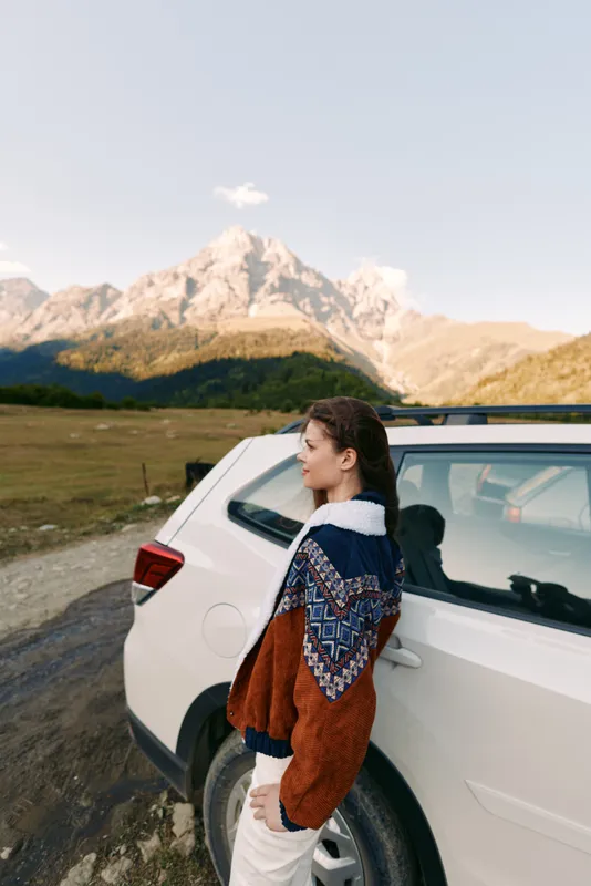Woman leaning against white car with cozy sweater in mountain landscape, outdoor travel and roadtrip portrait capturing scenic autumn adventure, peaceful nature and quiet exploration outdoors.