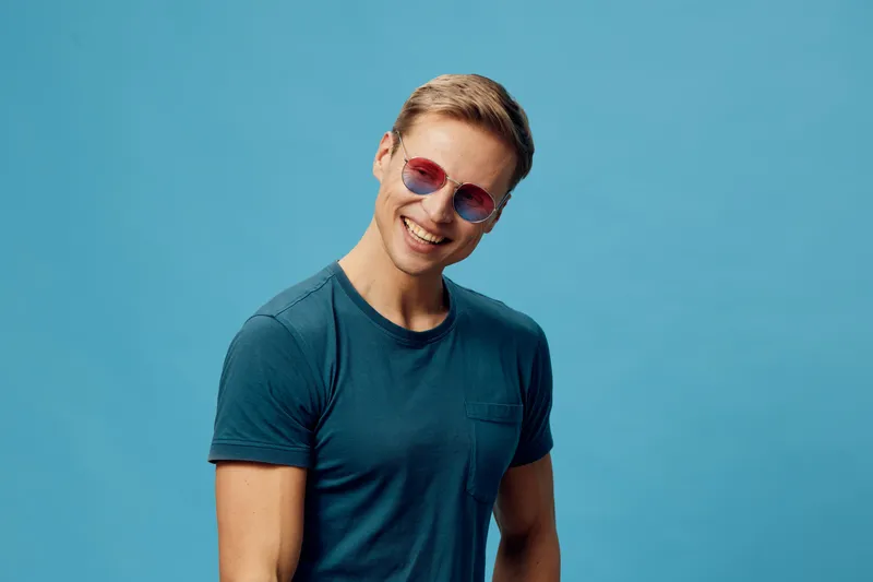 Smiling man with sunglasses and blue t-shirt against plain blue background holding mobile phone for lifestyle and leisure concept, cheerful casual male portrait, summer vibe, young adult, fashion styl