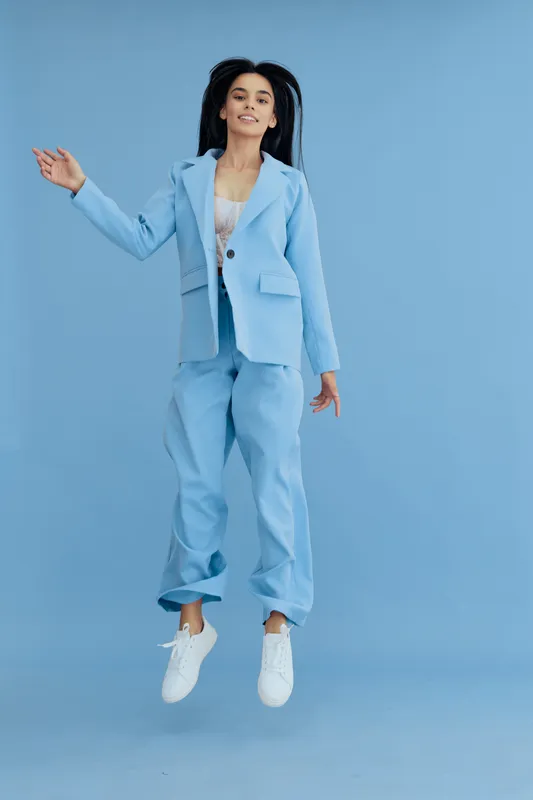 Young woman jumping wearing a light blue suit and white sneakers against a plain blue background, expressing energy and joy with a casual modern style.