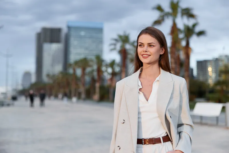 confident woman walking in light blazer on urban promenade with palm trees and modern buildings in soft evening light
