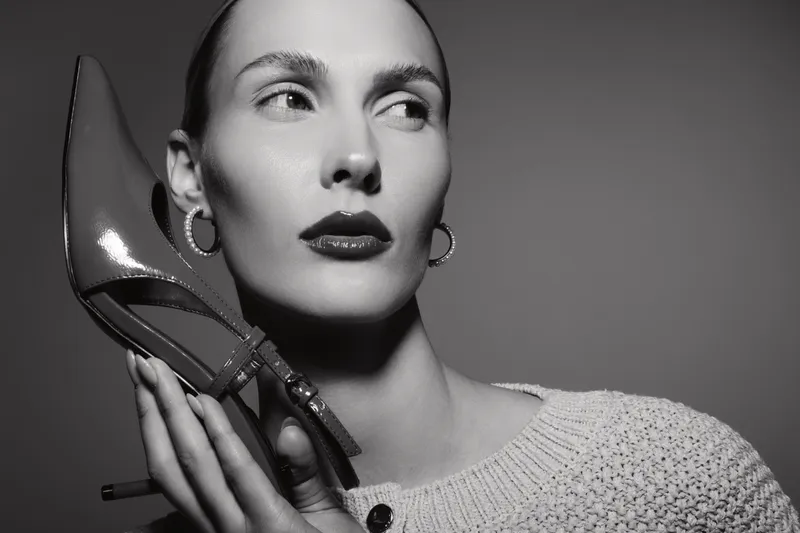 Elegant caucasian woman with pale skin holds shiny high heel shoe near face in black and white studio portrait. Fashion concept with confident female model and minimalistic background.