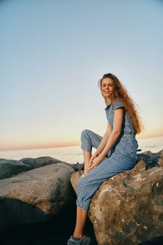 smiling woman sitting on rocks near ocean at sunset wearing denim jumpsuit with long curly hair and relaxed expression