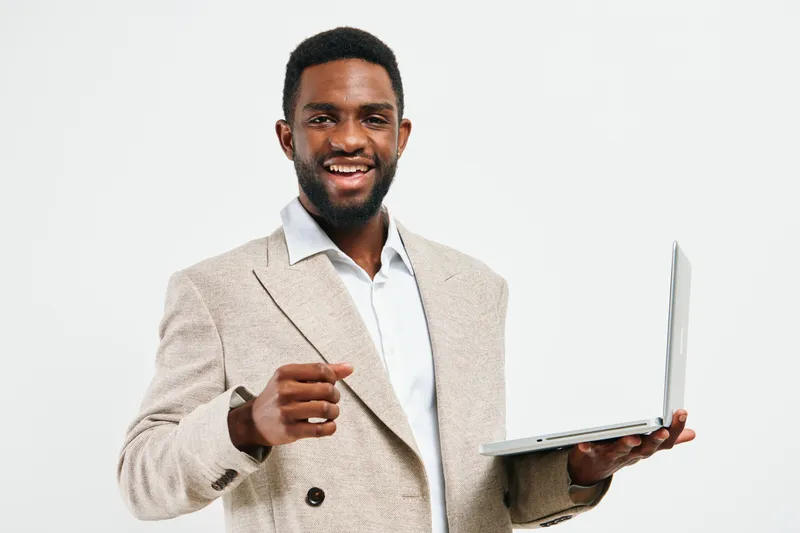 Smiling man with dark skin in business casual outfit holding laptop, gesturing confidently, on white background. Studio portrait of a cheerful professional male demonstrating success and positivity in