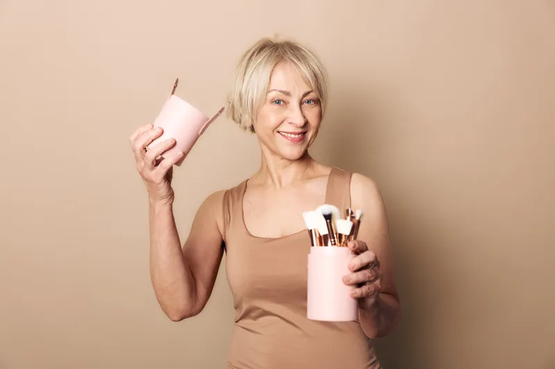 Smiling middle aged woman holding pink container with makeup brushes in one hand and lid in other, wearing beige tank top. Concept of beauty tools, cosmetics, and joyful lifestyle.