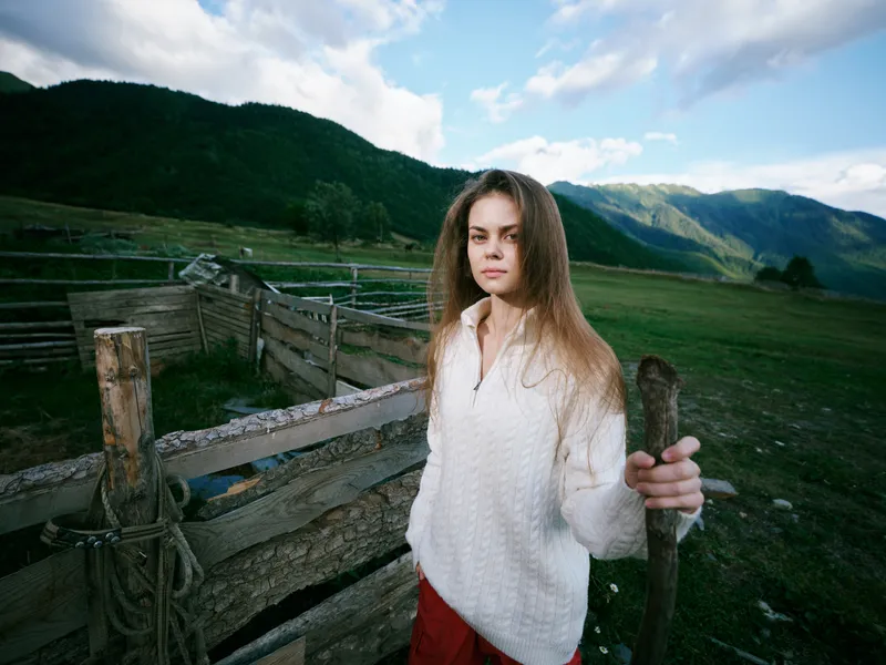 Elegant, casual woman standing beside a weathered wooden fence in a wide countryside, with green pastures, distant mountains, blue sky, and a tranquil, sunlit rural atmosphere