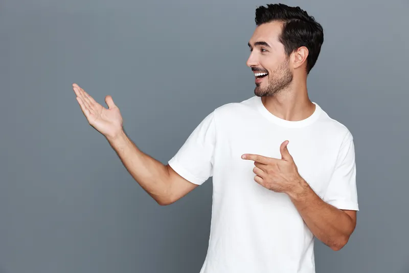 Smiling young man in casual white t-shirt gesture with open hand, look in camera and share enthusiasm isolated on solid gray background studio portrait. People lifestyle concept.