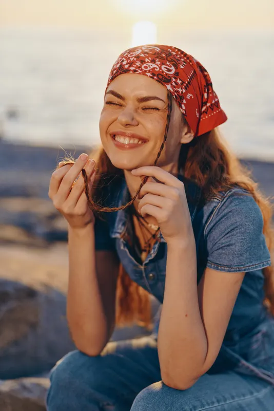 smiling woman wearing red bandana enjoying sunny outdoor moment with sunglasses and casual denim outfit near water during warm season