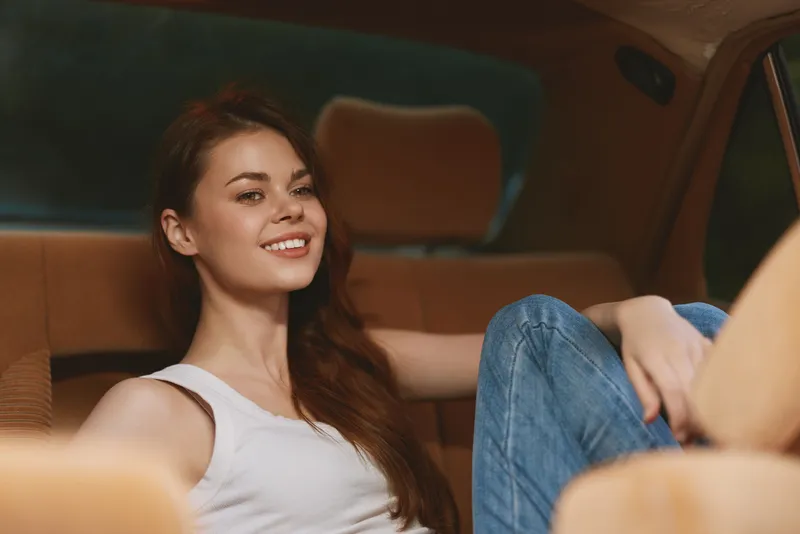 Young woman relaxing in a car, wearing a white tank top and denim jeans, smiling joyfully. The warm, neutral tones create a cozy atmosphere.